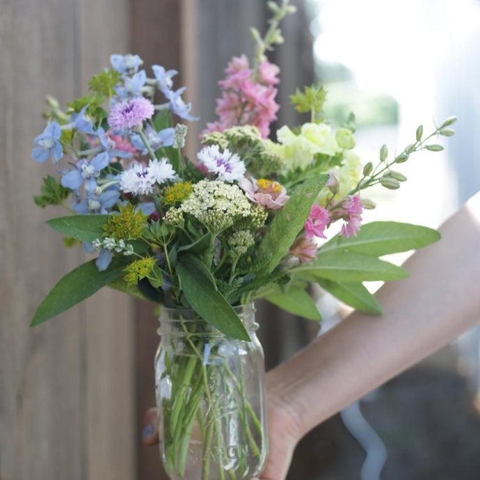 Hand holding a glass jar with a bouquet of colorful flowers against a blurred background